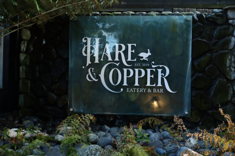 Hare & Copper Eatery & Bar glass sign, est. 2019, on a river-stone wall framed by ferns